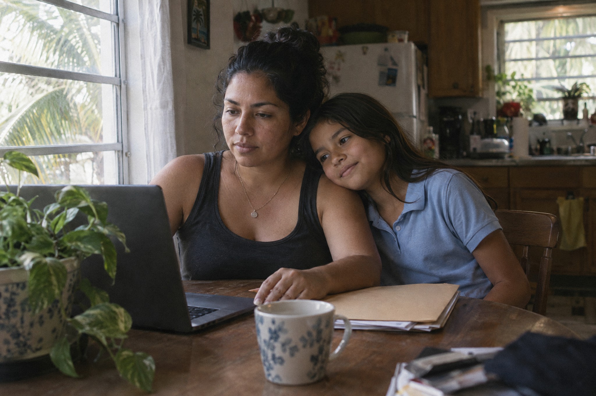 A mother reviews information on a laptop at the kitchen table while her daughter leans on her shoulder.